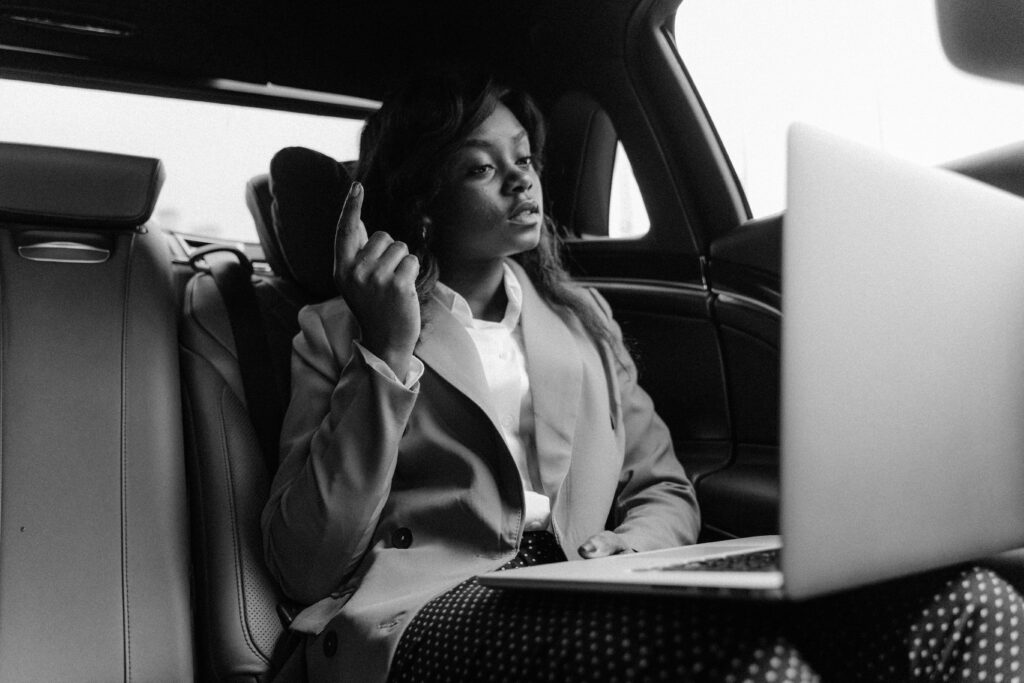 Black and white photo of a businesswoman working on a laptop in the car's backseat, poised and professional.